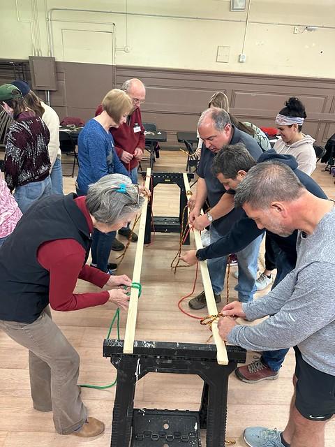 Group of adults learning practical knot skills, bent over a piece of wood propped up by sawhorses